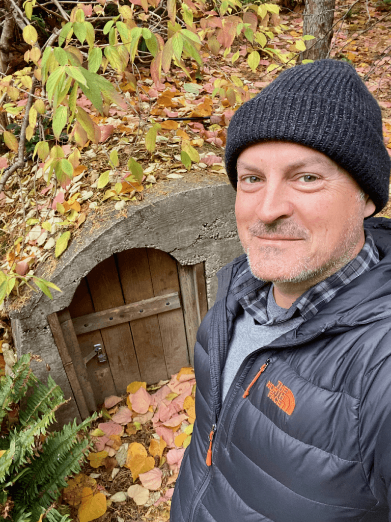 Matthew Kessi poses for a selfie in front of the hobbit door at Bellevue Botanical Garden. The leaves are changing to fall colors and dropping all around him. He's wearing a gray hat and jacket with orange accents.