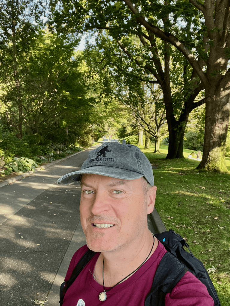 Matthew Kessi poses for a selfie at the Carl English Botanical Garden in Seattle. He's smiling and wearing a gray cap and magenta shirt. The trees behind him are a lush green and the roadway concrete gray.