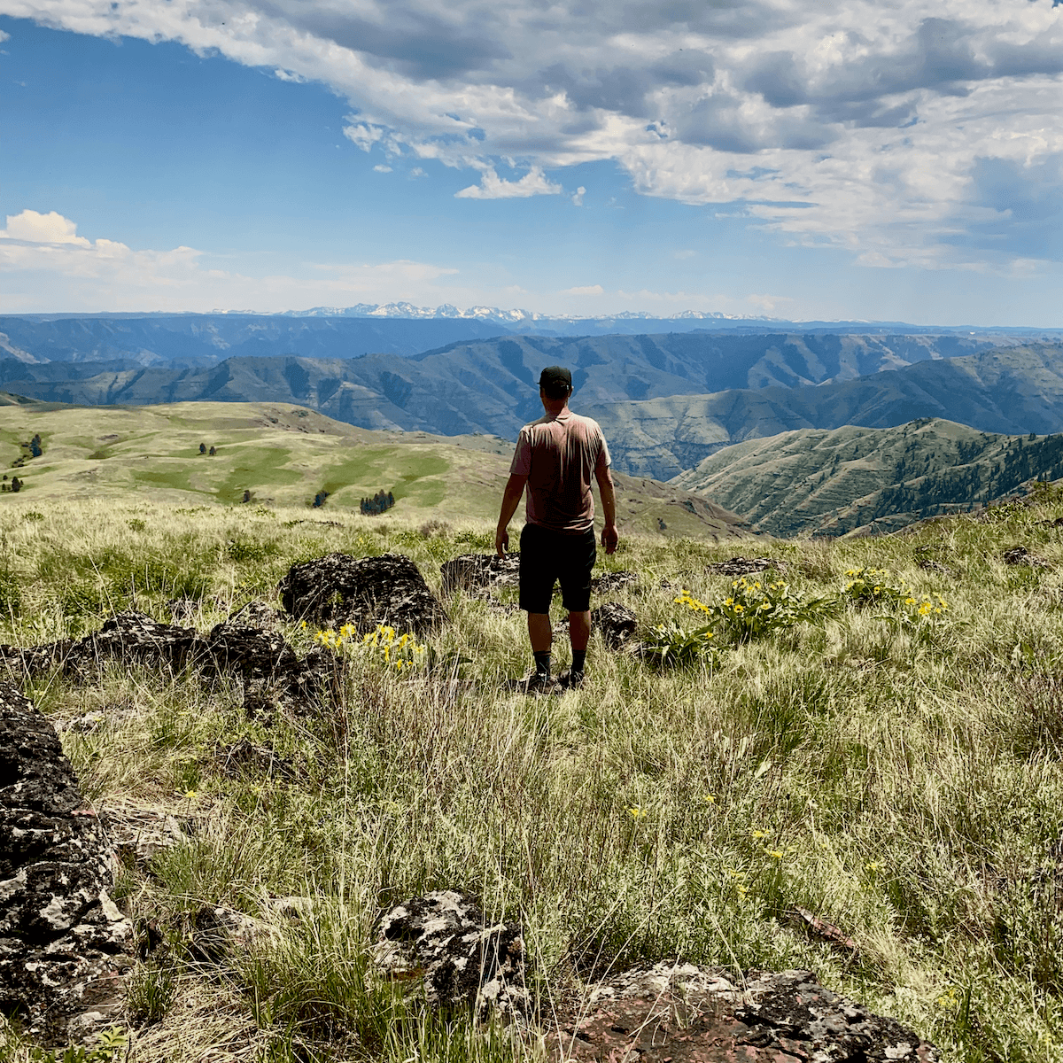 Matthew Kessi stands in his nature connection with a prairie that leads to dramatic green lined canyons toward mountains in the distance.