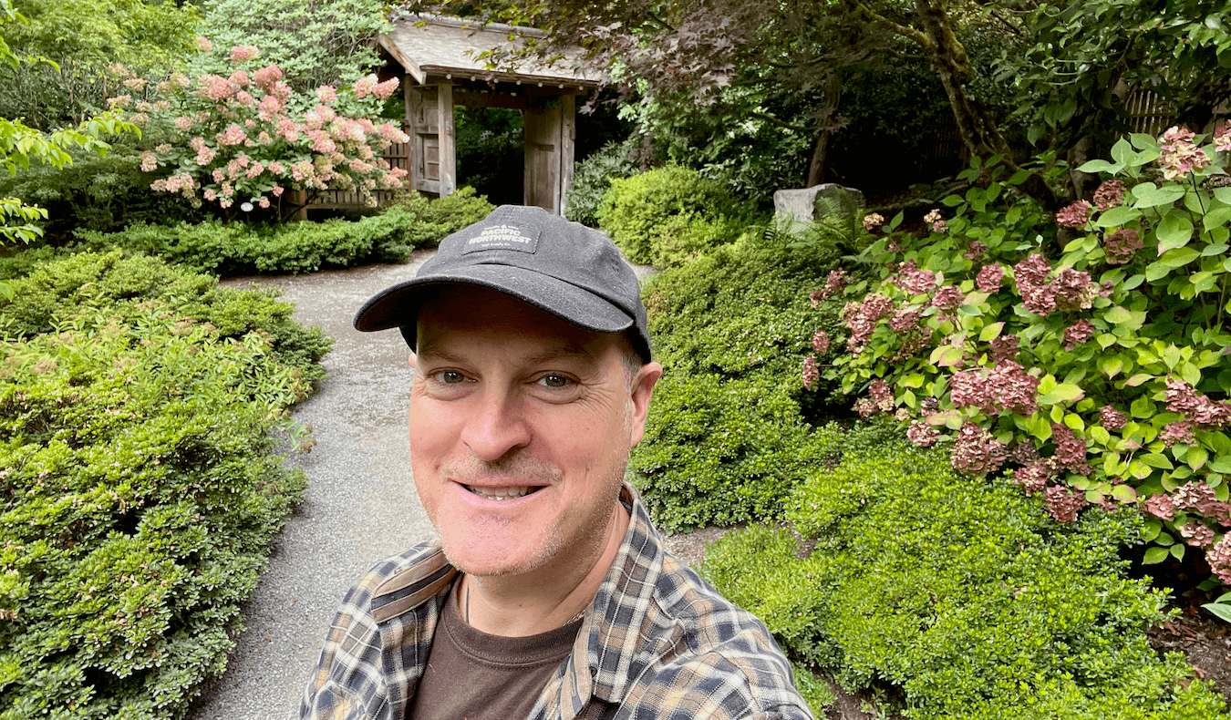 Matthew Kessi poses for a selfie in a Japanese themed garden. He's smiling and wearing a black cap and a brown plaid shirt. The bushes around him are green with hints of purple blooms fading.