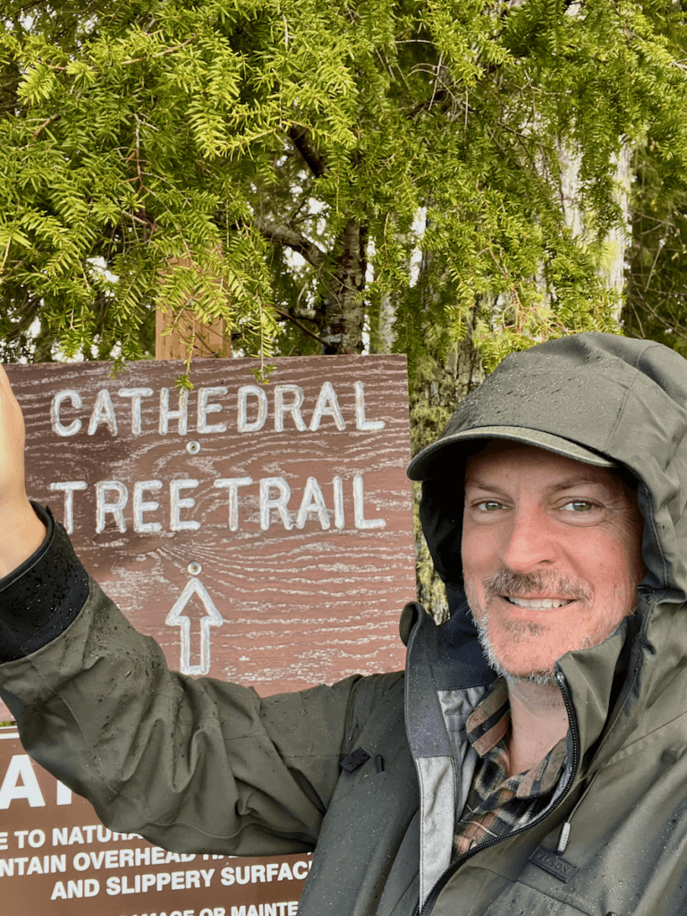 Matthew Kessi takes a selfie smiling next to a sign that says Cathedral Tree Trail, near the Astoria Column on the Oregon Coast.