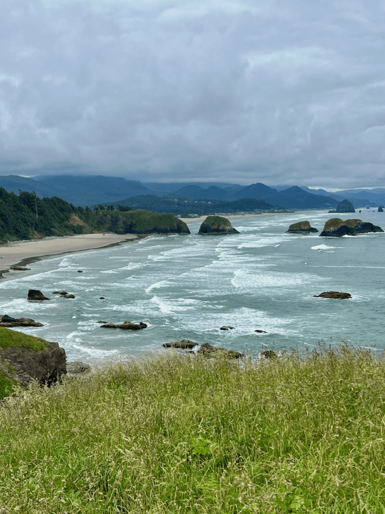 A photo of the Oregon Coast from iconic Ecola State Park near Cannon Beach. The waves majestically wash into a wide sandy beach while the gray and blue of the sky swirls together.