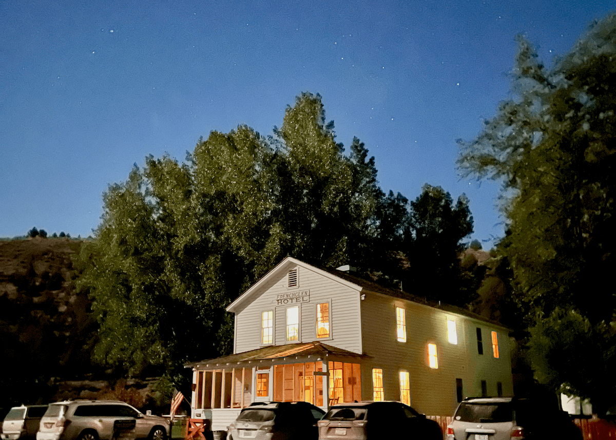 The Frenchglen hotel lights up the night with several windows shining bright as the stars in the sky above push out their light too. There are many cars parked in the front of the hotel, which is hidden under the brushy popular and cottonwood trees.