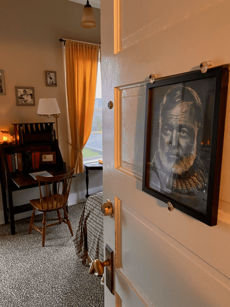 A photograph of Earnest Hemingway is attached to an old style door in the Silvia Beach Hotel in Newport Oregon. inside the room you can see an old fashioned wood desk and chair while orange curtains drape over a window.
