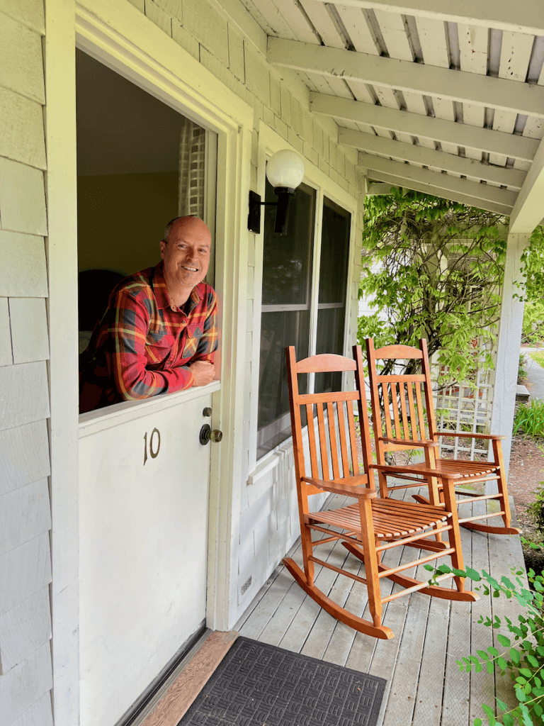 Matthew Kessi poses on the doorway of one of his favorite oregon and washington hotels, Lake Crescent Lodge. He's wearing an orange shirt and smiling while two wooden rockers hold space on the front porch.