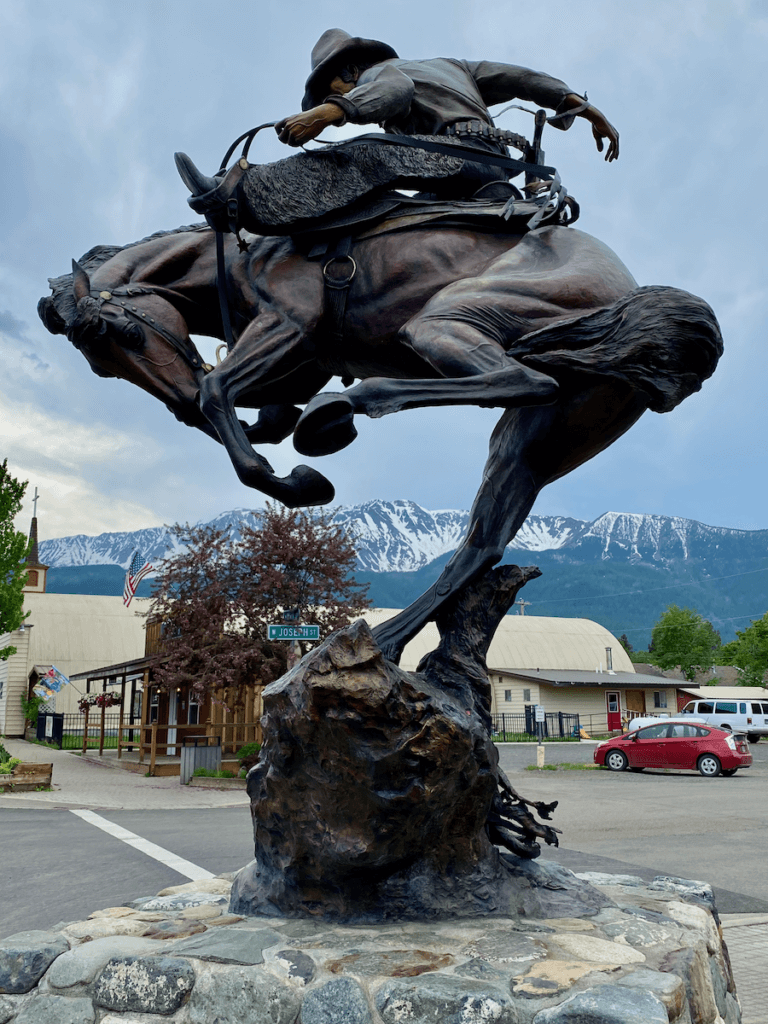 A statue of a cowboy riding a horse in bronze adorns the Main Street of Joseph, Oregon outside the Jennings Hotel. The Wallowa Mountains are dusted with snow in the background.