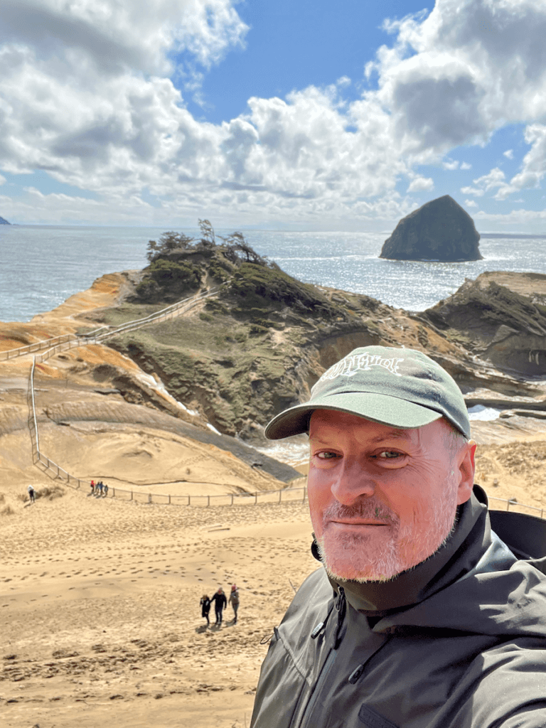 Matthew Kessi poses at Cape Kiwanda in Pacific City while hiking the Oregon Coast. He's wearing a green ball cap and a waterproof jacket. There are three hikers way below on the sand dune. Haystack rock is floating in the water in the ocean.
