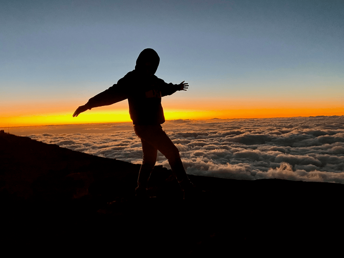 Matthew Kessi stands on the top of Haleakala Crater on Maui with the sun setting behind him. His silhouette is backlit by the orange glow of the sunset.