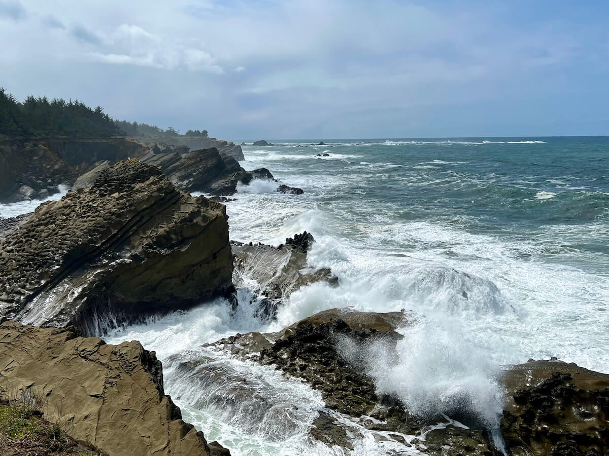 Waves crash along the path on a famous Oregon Coast Hike at Shore Acres State Park. The sky is a swirl of gray and blue and the water foamy as it crashes on the rock formations.