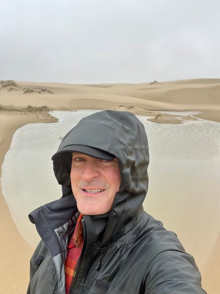 Matthew Kessi poses for a selfie while hiking on the Oregon Dunes. There is a pool of water behind him and more dunes continuing into the horizon.
