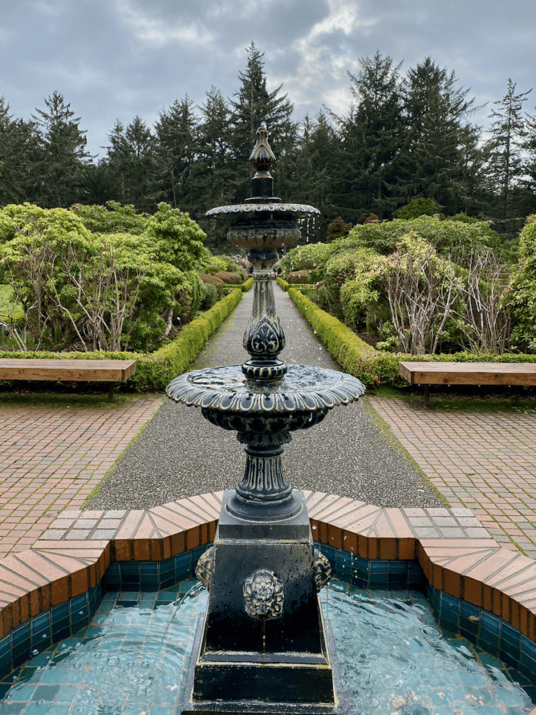 An ornate fountain drips water into a blue pool while surrounded by a brick pathway. There are shrubs leading down a walkway toward a forest of evergreen trees.