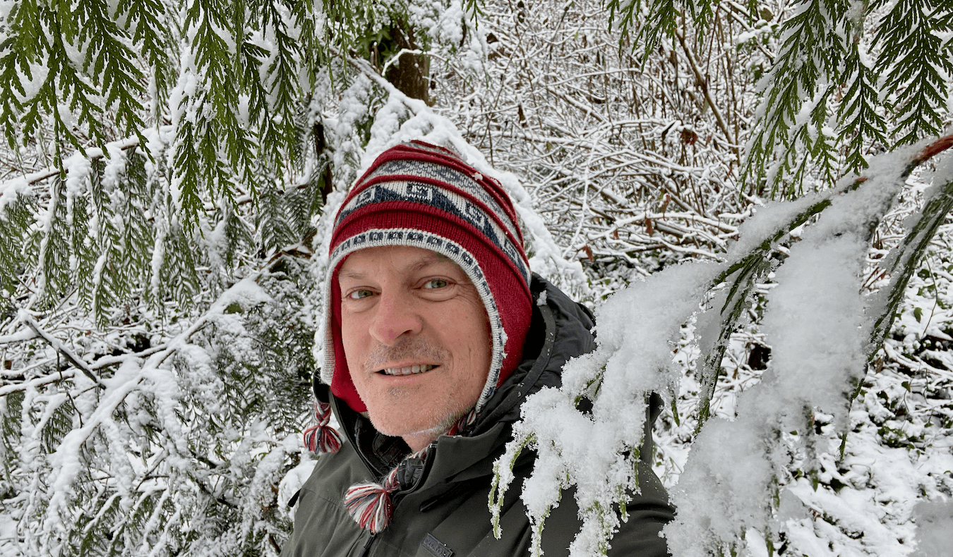 Matthew Kessi poses for a selfie in the middle of woods with snow dripping over cedar leaves and onto bushes in the background. He is smiling while wearing a red cap.