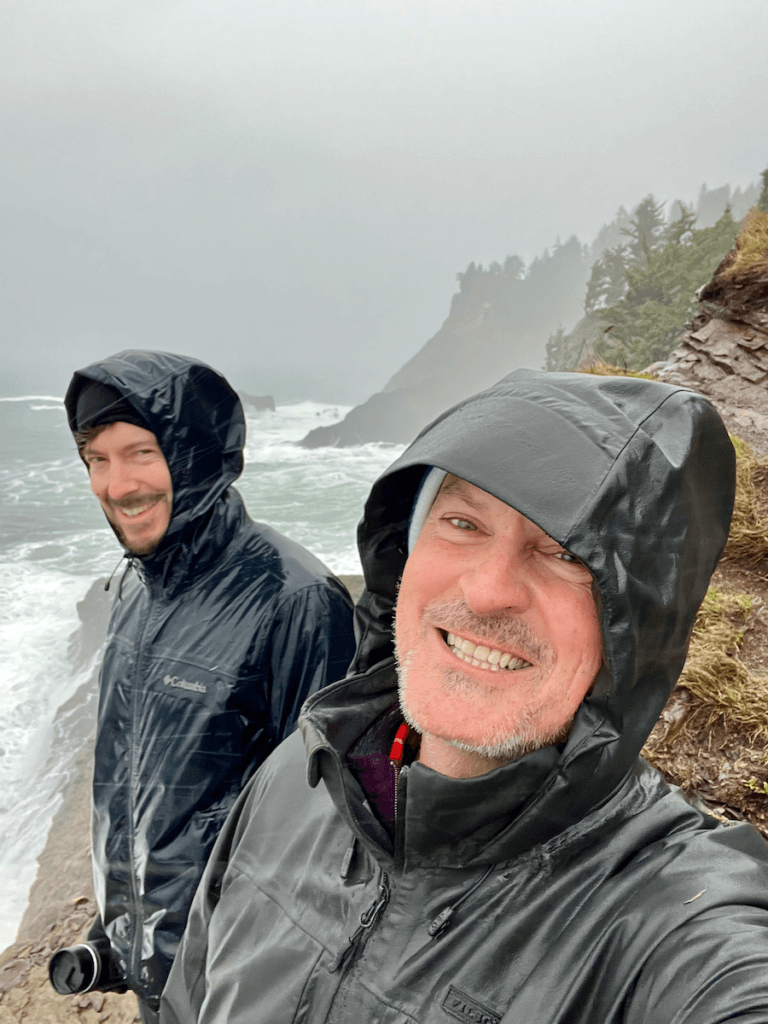 Matthew Kessi and Brian Turner pose for a selfie at the end of Cape Falcon Trail on the Oregon Coast. They are both in raingear smiling because they are very wet. In the background waves crash against rock stacks at Oswald West State Park.