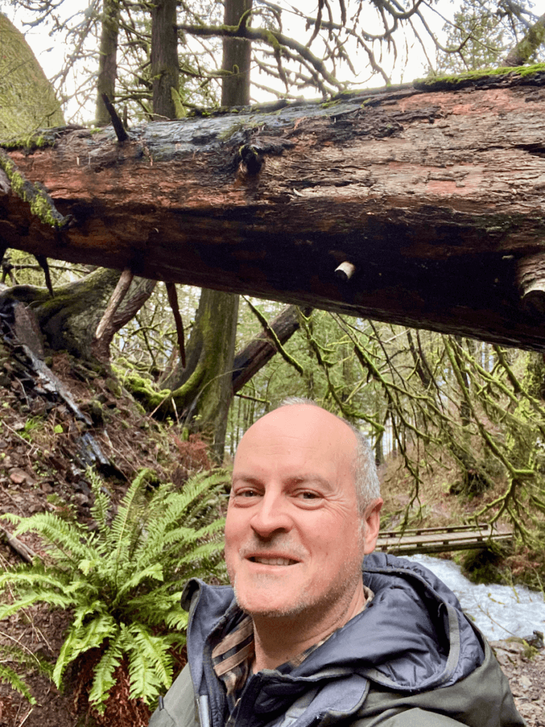 Matthew Kessi smiles while on a forest bathing hike in the Pacific Northwest. He's underneath a giant fir log that has fallen high above a trail. In the background a rushing creek passes under a footbridge.