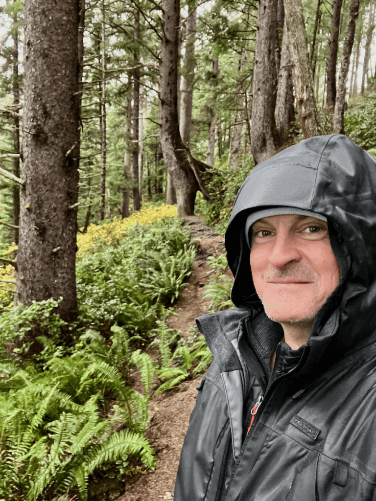 Matthew Kessi poses for a selfie while forest bathing in Seattle. He's wearing a dark green rain jacket and smiling. Behind him are ferns and spruce trees.