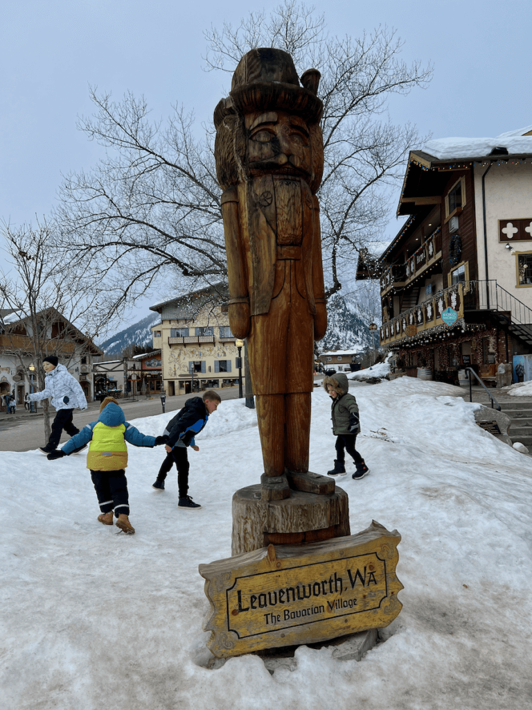 A wood carved nutcracker pops out of snow in the downtown center of Leavenworth Washington. In the background young children are playing in the snow.