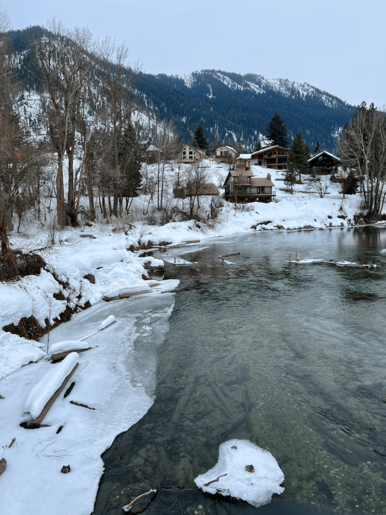 The Wenatchee River flows through a snowy winter landscape in Leavenworth, Washington. There are chalet homes scattered among the trees and mountains.