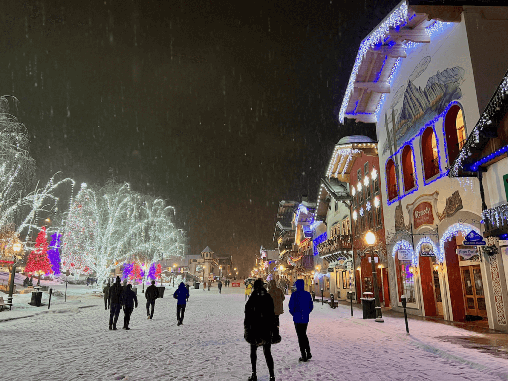 People wander in the snow as it falls on the front street of Leavenworth, Washington. The buildings are lit up in this evening shot and you can see the snowflakes falling.