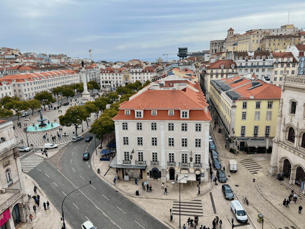 Downtown Lisbon is abuzz with pedestrians walking in the square below. This Birdseye view showcases the orange tile roofs of the buildings.