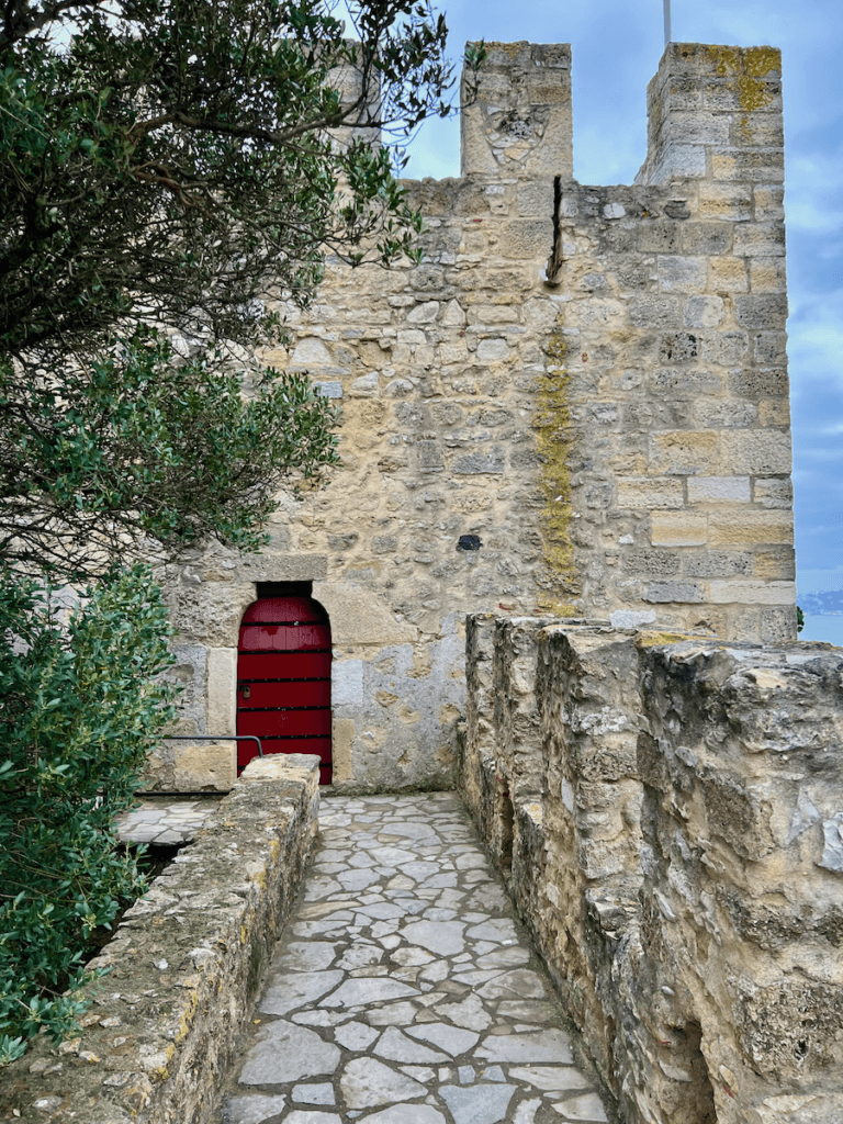 The castello de S. Jorge is a dramatic bastion at the top of a hill in Lisbon Portugal. The ramparts are seen here leading to a bright red door framed in by an ancient olive tree and blue skies. This is a favorite nature forward thing to do while on vacation in Portugal.