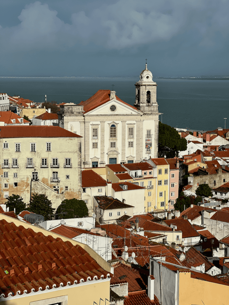 A cathedral rises above the orange roofs of Lisbon Portugal under gray stormy skies.