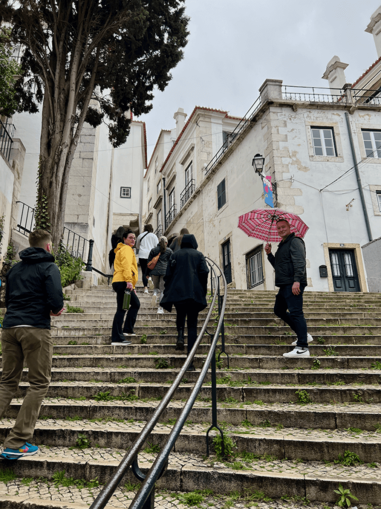 A group of people on a nature forward vacation walk up the many steps to the top of a hill in Lisbon Portugal. A man looks back at the camera holding a red plaid umbrella as a lady in yellow rain jacket turns toward the camera too. The buildings in the background are white brick under gray skies.