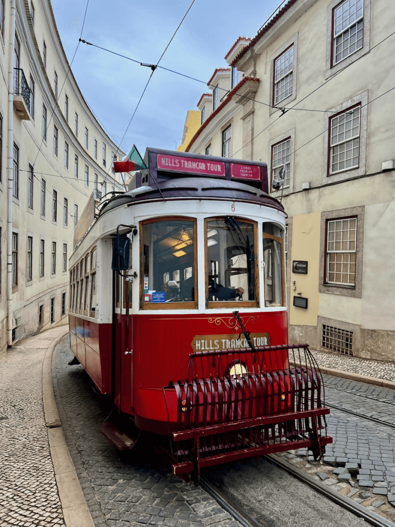 A cable car in Lisbon Portugal moves along a cobblestone street. It is bright red and attached to wires from above.