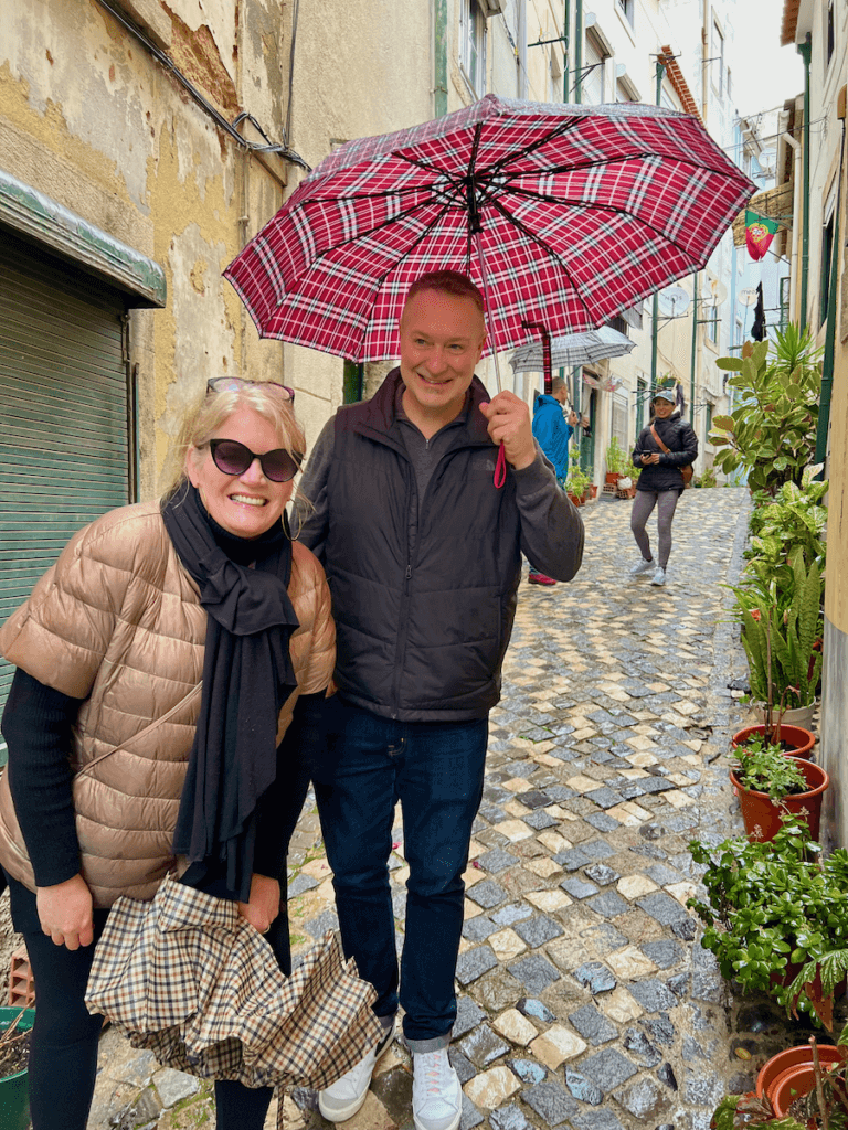 A man and woman are smiling under a red plaid umbrella while on a nature-forward walking tour of Lisbon Portugal. The cobblestone street they're walking has a line of plants in pots against an orange washed wall.