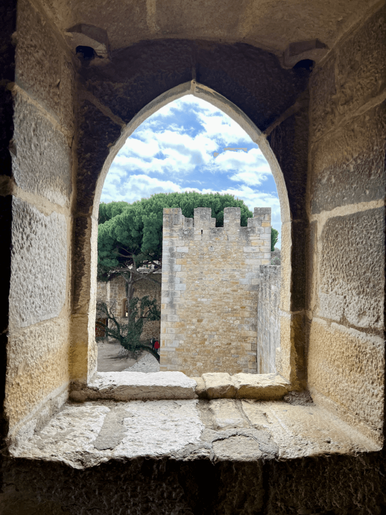 The Castelo de St. Jorge in Lisbon Portugal taken from the inside of the castle. The bricks and mortar can be seen in the foreground while in the background a large olive tree can be seen under blue sky with puffy white clouds.