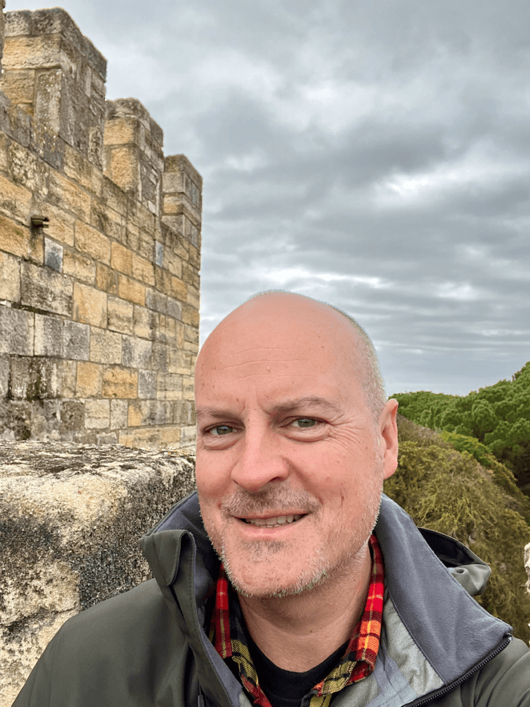 Matthew Kessi poses for a selfie at the Castelo de St. Jorge atop Lisbon Portugal. He's smiling and behind him are the ramparts of the castle as well as a dense canopy of green trees. The sky in the background is gray.