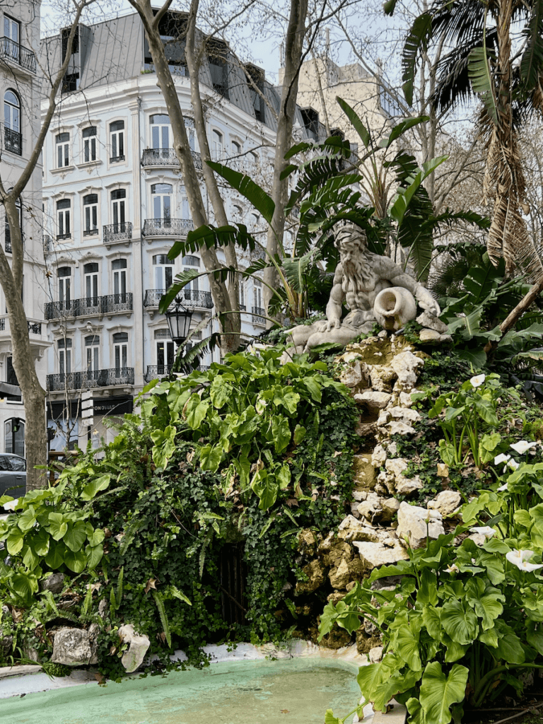 A sculpture of a stone man with a spilling jug of water hails over a pond below. He's surrounded by assorted green leafy plants. In the background is an ornate building along a fashionable boulevard in Lisbon Portugal.