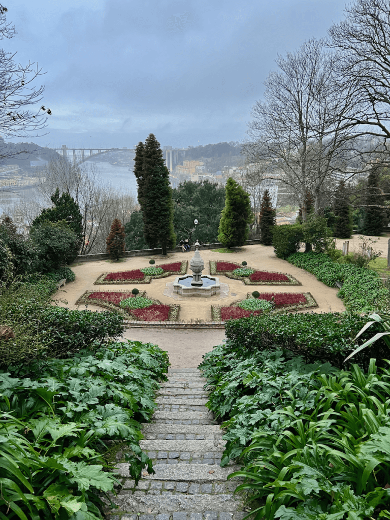 A formal garden in Porto Portugal shows of a four flower bed combination with green textures and magenta ground cover around an ornate fountain. The Douro River flows in the distance.