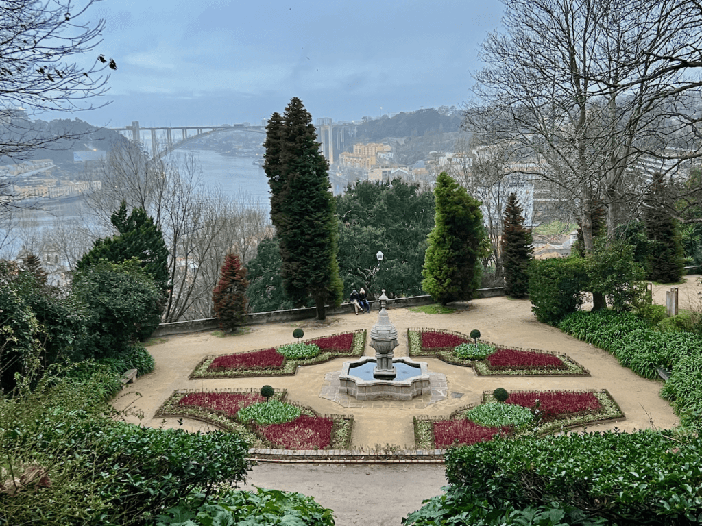 A formal garden in Porto Portugal shows of a four flower bed combination with green textures and magenta ground cover around an ornate fountain. The Douro River flows in the distance.