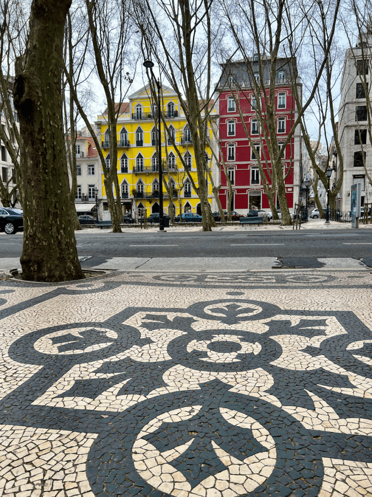 A yellow and red building stand proudly on the other side of a Wide boulevard in downtown Lisbon Portugal. The trees in between are without their leaves because it is still winter. In the foreground is a beautiful black and white mosaic on the sidewalk.