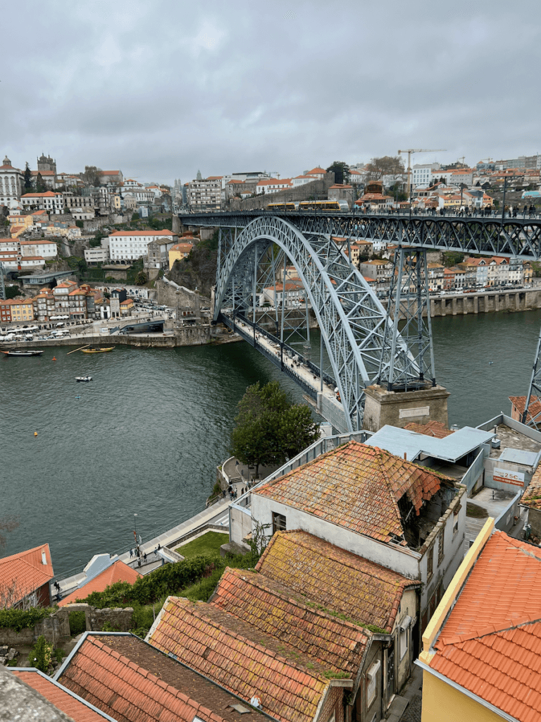 A metal bridge spans a river in Porto Portugal. Many layers of houses with orange tiled roofs can be seen.