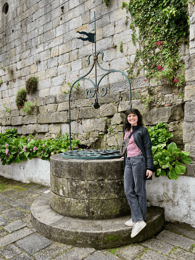 A young woman stands near a well in a courtyard of a church in Porto, Portugal. She is smiling wide and surrounded by green garden plants.