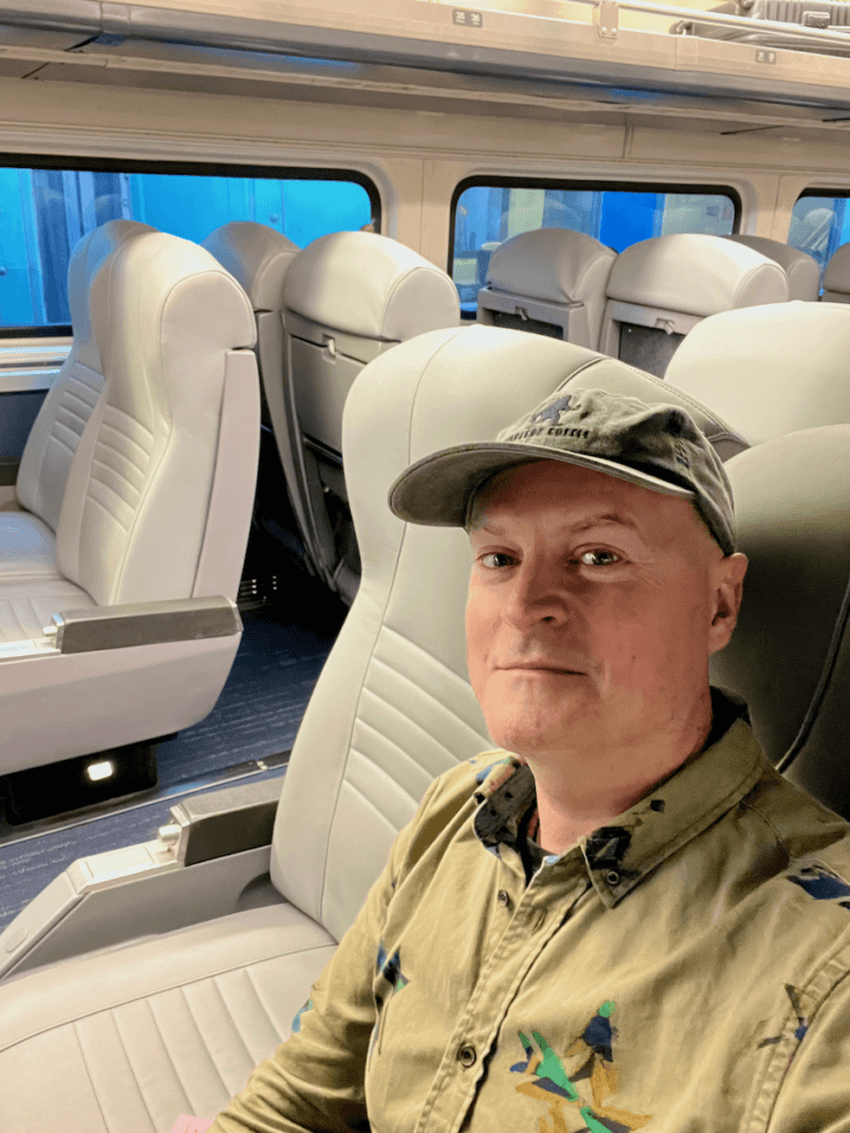 Matthew Kessi poses for a selfie in a car on an Amtrak train. He is sitting in plush gray leather seat with others visible behind him. He's smiling and wearing a ball cap and a tan shirt with block prints.