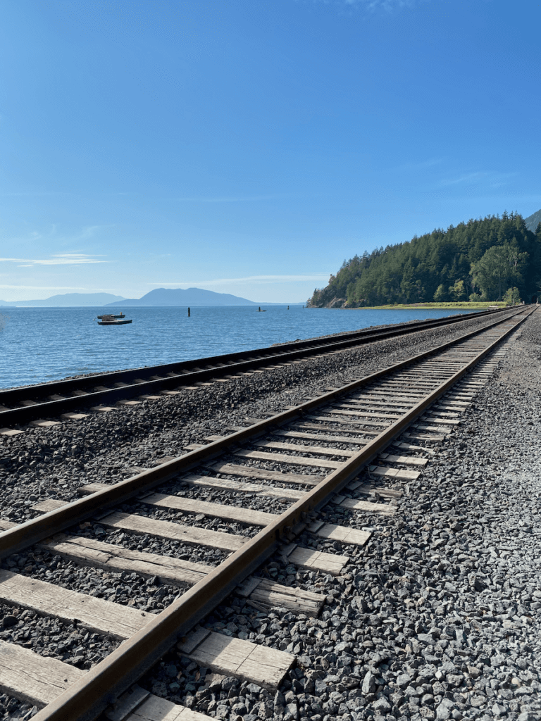 Railroad tracks pass by the blue waters of the Salish Sea used by Amtrak on trains from Seattle. In the background you can see the San Juan Islands. The sky is bright blue and land along the tracks has lime green grass and darker green trees.