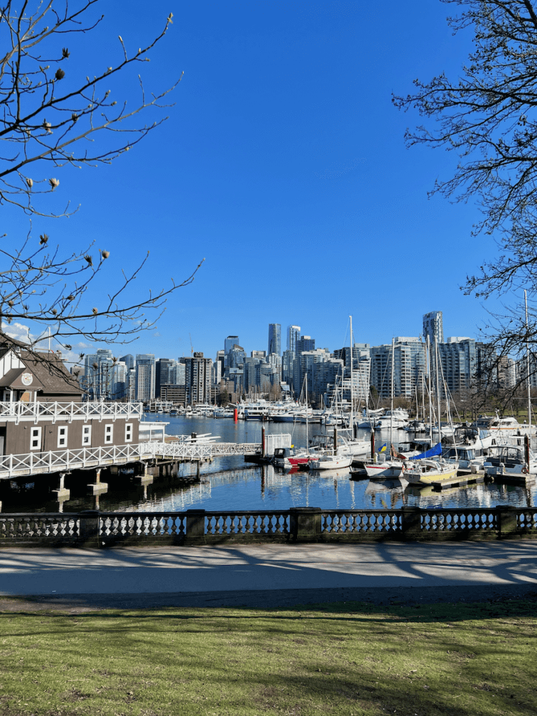 Downtown Vancouver Canada in the blue sky sunshine is beautiful. Here a historic boathouse with brown and white paint sits next to a marina with sailboats. In the distance you can see the high-rise skyling of downtown Vancouver, British Columbia.