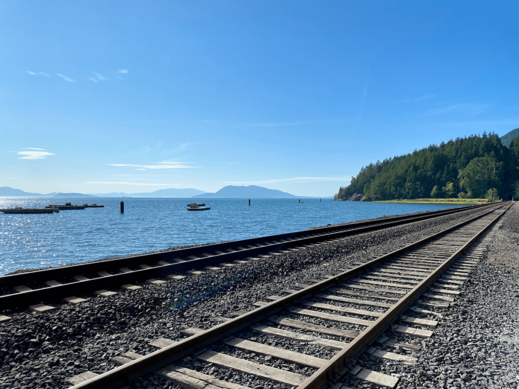 Railroad tracks pass by the blue waters of the Salish Sea. In the background you can see the San Juan Islands. The sky is bright blue and land along the tracks has lime green grass and darker green trees.