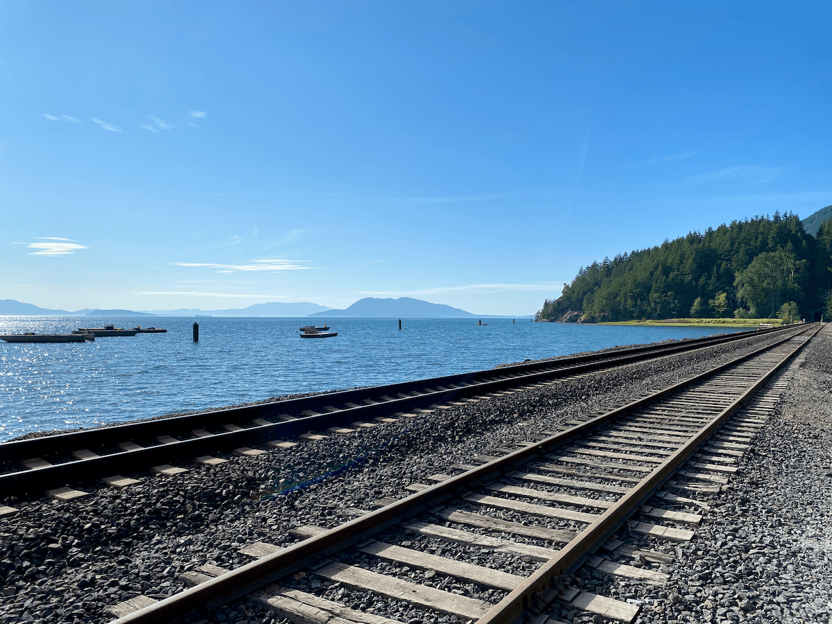Railroad tracks pass by the blue waters of the Salish Sea. In the background you can see the San Juan Islands. The sky is bright blue and land along the tracks has lime green grass and darker green trees.