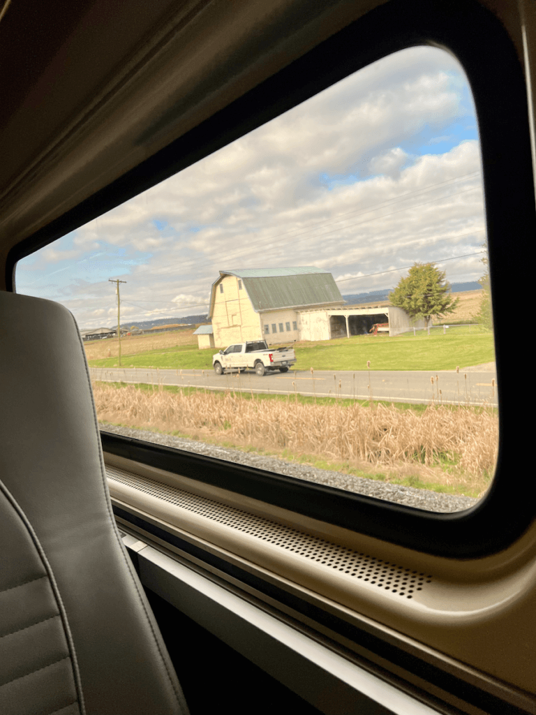 A view of the scenery from the train that rides between Vancouver and Seattle. The edge of the leather seat is visible and outside the window a truck rides along a country road in front of a historic looking white barn under blue sky with clouds.