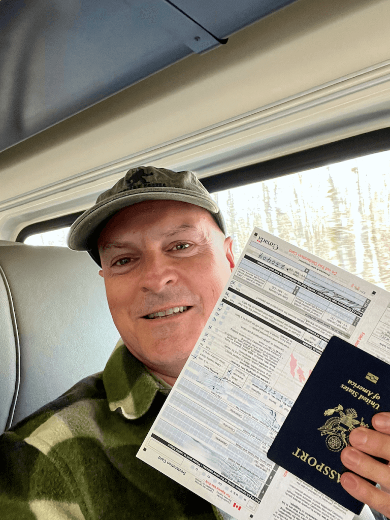 Matthew Kessi poses for a selfie with his passport and a Canadian Customs form in preparation for entering Canada. He is wearing a ball cap and a green plaid coat.