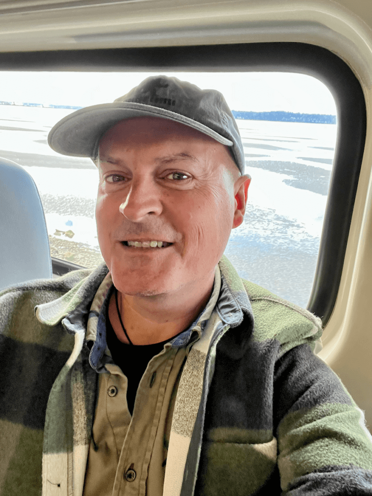 Matthew Kessi poses for selfie on the train from Seattle to Vancouver. He's smiling while wearing a ball cap and a few layers of green clothing. In the background you can see the tidal flats of the Salish Sea.