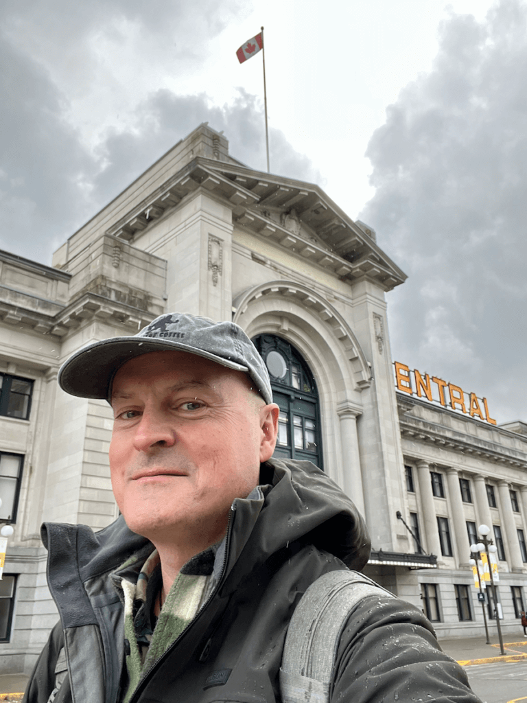 Matthew Kessi poses for a selfie in front of the Pacific Central train station in Vancouver Canada. The canadian flag is waiving high above the statuesque stone building.