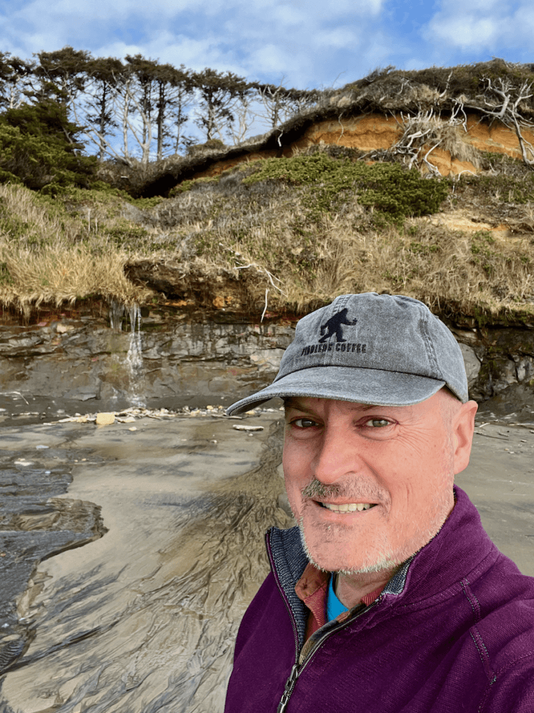 Matthew Kessi poses for a selfie on a one day trip to Newport Oregon. He's wearing a gray cap and purple sweater and smiling. Behind him you can see layers of earth leading up to a forest of weathered coastal trees.