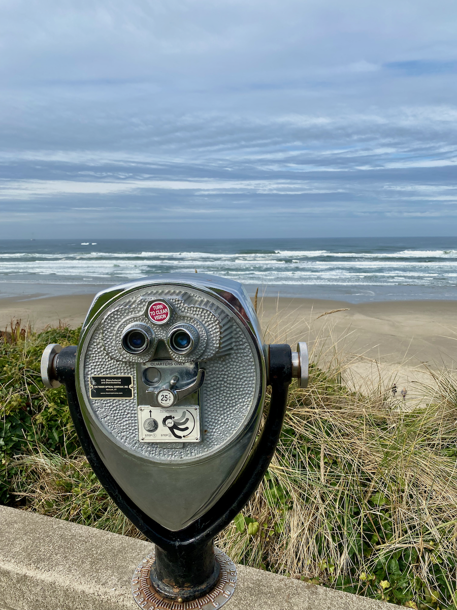 A shiny silver binocular is attached to a wall looking over to the flat sands of Nye Beach in Newport, Oregon. The waves are churning in the surf with a gray sky above.