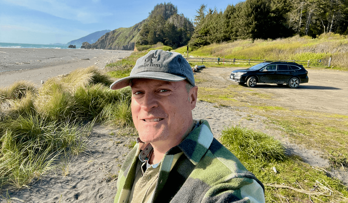 Matthew Kessi poses for a selfie at a secluded beach on the Oregon Coast. A subaru is parked in the lonely lot and he's smiling and wearing a gray cap and a green plaid coat. The beach and waves of the Pacific Ocean are crashing in the distance.