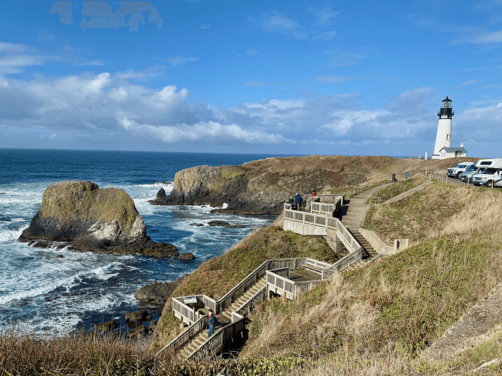 Yaquina Head Lighthouse rises up on a spit of land surrounded by waves crashing against dramatic rock stacks. There are some cars parked right on the edge of the cliff with a staircase leading down to the rocky beach.