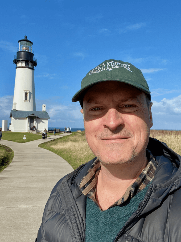 Matthew Kessi poses for a selfie outside Yaquina Head LIghthouse in Newport Oregon. He's wearing a green cap and smiling under a bright blue sky.